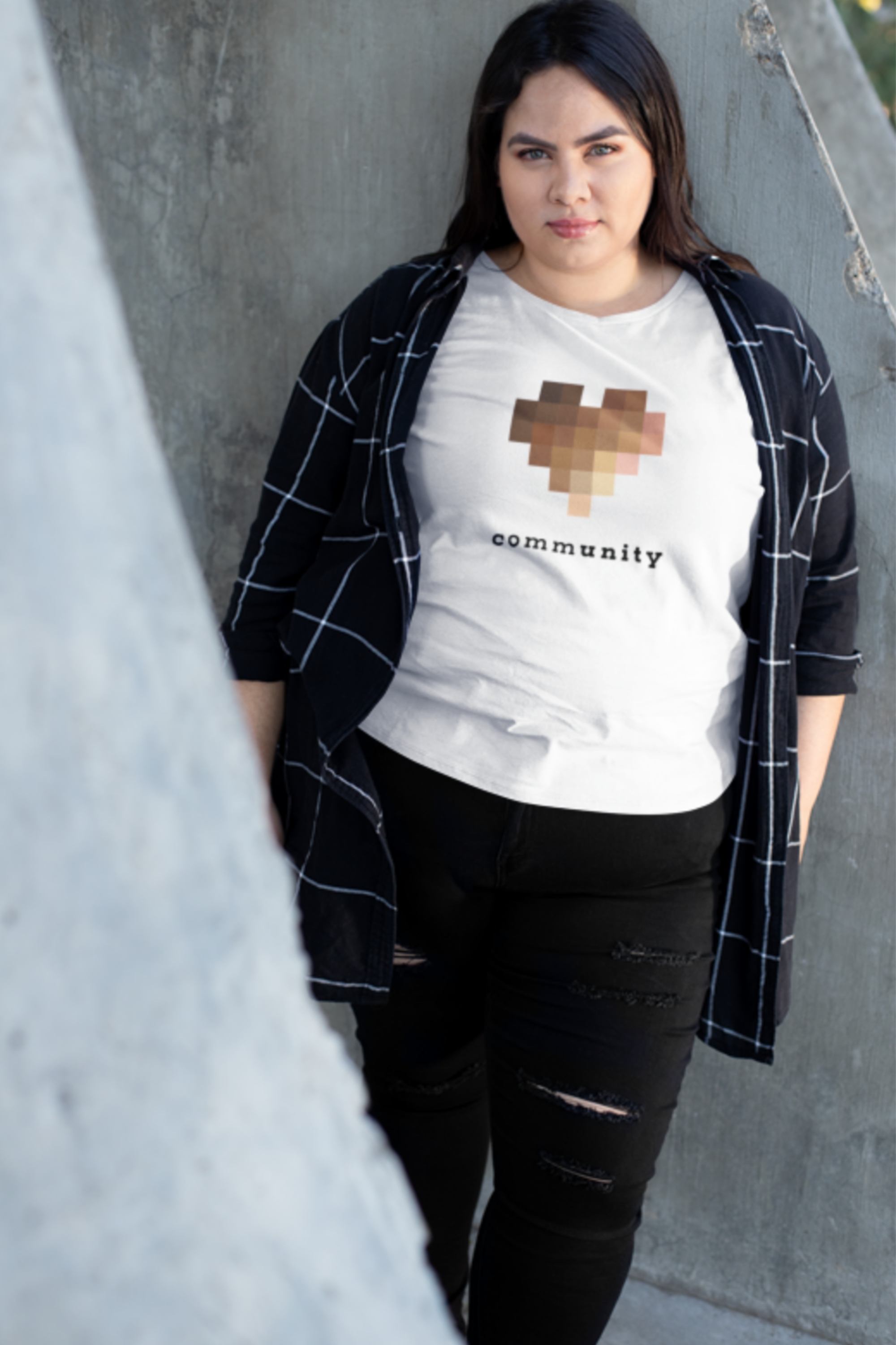 A female presenting model is wearing a solid white unisex, crew-neck t-shirt while leaning on a concrete wall.  The central graphic on the chest is a large pixelated heart composed of blurred squares in varying skin-tone shades. Below the heart, the word "community" is printed in lowercase letters. The font alternates between bold and light weights, highlighting the letters "unity."
